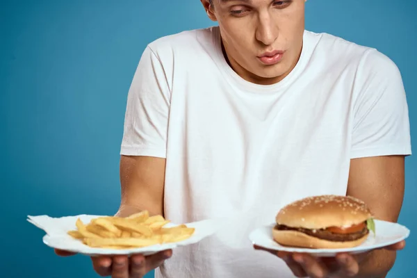 Junge Kerl mit Pommes und Hamburger auf blauem Hintergrund interessiert Blick Emotionen Fast Food Kalorien beschnitten Ansicht Copy Space — Stockfoto