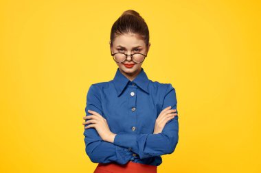 Gözlüklü ve mavi gömlekli kadın sarı arka planda el kol hareketi yapıyor.