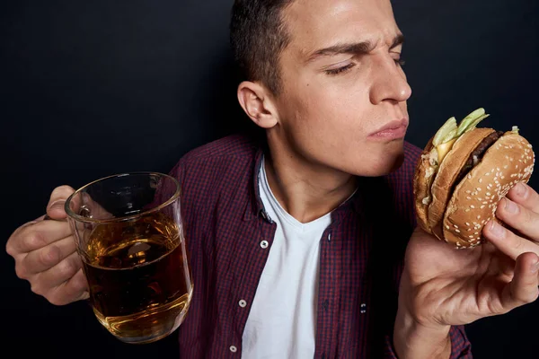 Hombre con cerveza taza y hamburguesa en manos divertido estilo de vida estudio oscuro aislado fondo — Foto de Stock