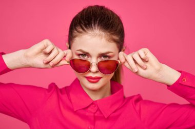 Pembe tişörtlü, elinde gözlüklü, arka planda izole edilmiş manzaralı moda kadını.