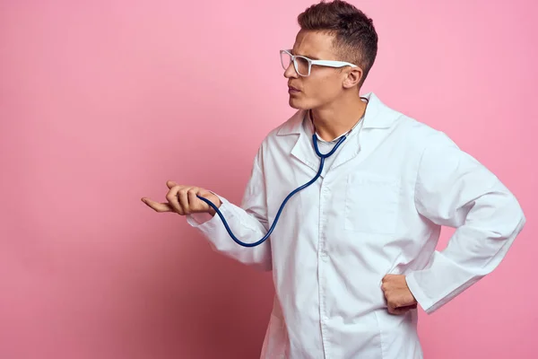 Male doctor in a medical gown with a stethoscope around his neck on a pink background and glasses on his face — Stock Photo, Image