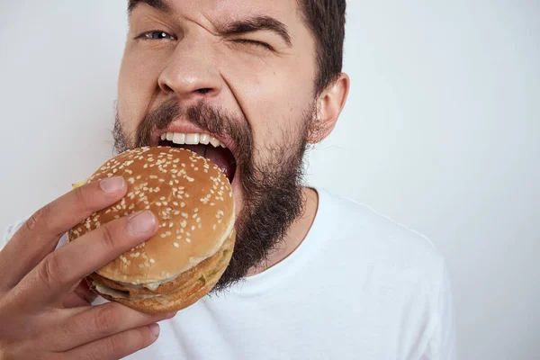 Ein Mann, der einen Hamburger auf hellem Hintergrund in einem weißen T-Shirt isst, hat die Nahaufnahme von Hunger-Fast Food abgeschnitten — Stockfoto