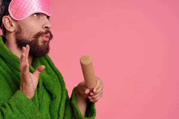 Um homem com uma máscara de dormir cor-de-rosa no rosto com um roupão verde com um rolo na mão — Fotografia de Stock