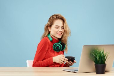 Dizüstü bilgisayar kulaklığının önündeki masada oturan bir kadın çevrimiçi yaşam tarzı mavi arka planda oynuyor.