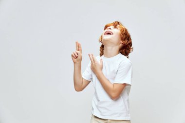 Neşeli kızıl saçlı bir çocuk yukarı bakar ve parmaklarıyla beyaz bir tişört gösterir.