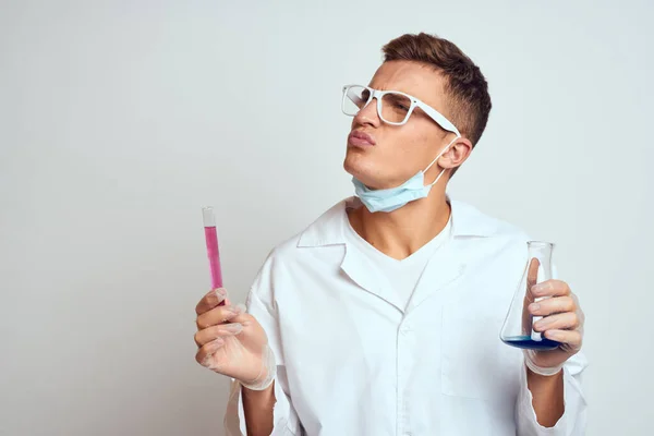 Un assistant de laboratoire en blouse médicale avec un masque de protection tient une fiole avec un liquide de réaction chimique — Photo