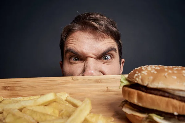 Hombre emocional con paleta de madera comida rápida hamburguesa papas fritas comer comida estilo de vida oscuro fondo — Foto de Stock