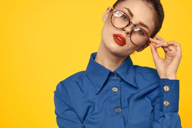 Mavi gömlekli, gözlüklü, yüzünde parlak makyajlı bir kadın elleriyle işaret ediyor.