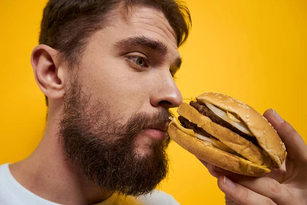 Hombre con hamburguesa en manos de comida rápida dieta blanca camiseta primer plano amarillo fondo — Foto de Stock