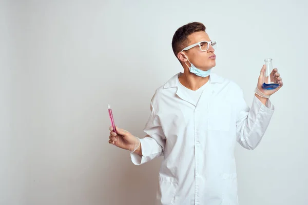 Un assistant de laboratoire en blouse médicale avec un masque de protection tient une fiole avec un liquide de réaction chimique — Photo