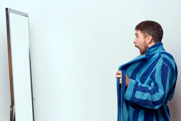 Een man in een blauw gewaad onderzoekt zichzelf in een spiegel in een heldere kamer bijgesneden uitzicht — Stockfoto