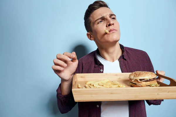 Hombre con paleta de madera comida rápida papas fritas hamburguesa hambre dieta alimentos fondo azul — Foto de Stock