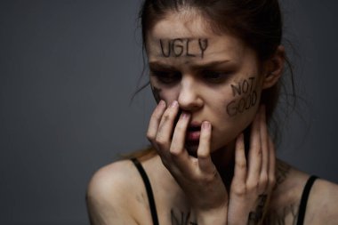 Hüsrana uğramış bir kadın vücudunda çirkin yazıtlar ve gri bir arka plan portresi üzerinde siyah bir tişört var. 