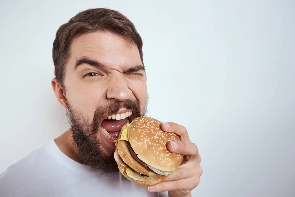 Beyaz bir tişörtle arka planda hamburger yiyen bir adam yakın çekim açlık ve fast food manzarası izledi. — Stok fotoğraf