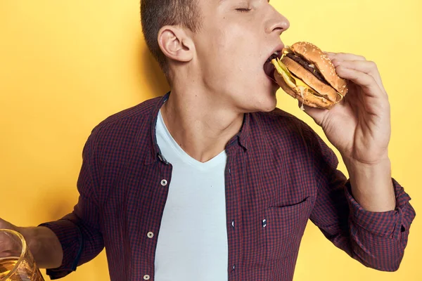 Homem alegre com uma caneca de cerveja e um hambúrguer em suas mãos dieta comida estilo de vida fundo amarelo — Fotografia de Stock