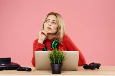 Kulaklık takan, dizüstü bilgisayarlı bir kadın masa başı eğlence sektöründe pembe arka planda oturuyor.