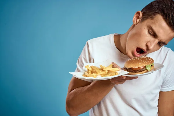 Jovem com batatas fritas e hambúrguer no fundo azul interessado olhar emoções fast food calorias corte vista Copiar Espaço — Fotografia de Stock