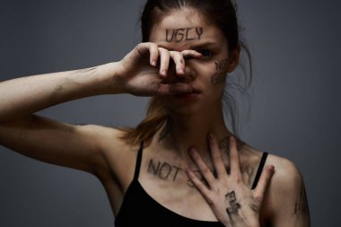 Hüsrana uğramış bir kadın vücudunda çirkin yazıtlar ve gri bir arka plan portresi üzerinde siyah bir tişört var. 