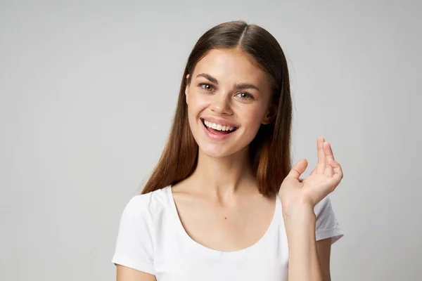 Γυναίκα με ένα εκθαμβωτικό χαμόγελο σε ένα γκρι φόντο σε ένα λευκό μπλουζάκι gesticulate με ένα χέρι — Φωτογραφία Αρχείου