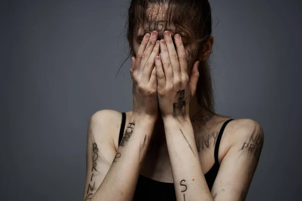 Засмучена жінка з образливим написанням на своєму тілі на сірому фоні, торкаючись себе обрізаним поглядом — стокове фото