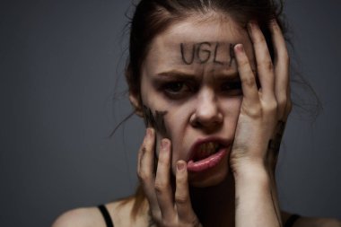 Gri arka planda kırıcı yazılar yazan üzgün kadın elleriyle kendine dokunuyor.