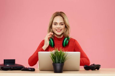 Kulaklık takan, dizüstü bilgisayarlı bir kadın masa başı eğlence sektöründe pembe arka planda oturuyor.