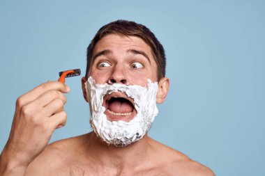 Yüzünde jilet ve beyaz köpük olan bir adam. Mavi arka plan çıplak gövde banyo prosedürleri.