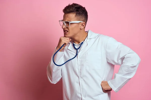 Male doctor in a medical gown with a stethoscope around his neck on a pink background and glasses on his face — Stock Photo, Image