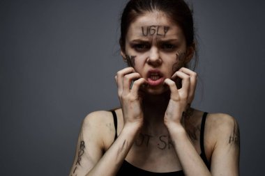 Hüsrana uğramış bir kadın vücudunda çirkin yazıtlar ve gri bir arka plan portresi üzerinde siyah bir tişört var. 