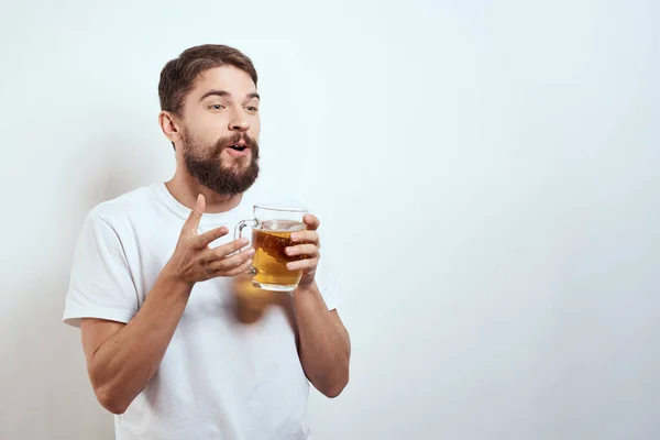 Hombre con una taza de cerveza en sus manos y una camiseta blanca fondo claro bigote barba emociones modelo — Foto de Stock