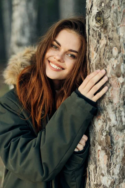 Ormanda sıcak ceketli neşeli bir kadın gülümseyerek bir ağaca yaslandı. — Stok fotoğraf