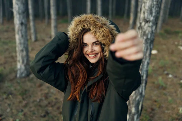 Uma mulher sorrindo em uma jaqueta de outono com um capuz em um fundo da floresta segura a mão na frente dela — Fotografia de Stock