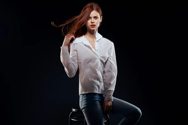 Красива жінка сидить на стільці позує білі сорочки джинси довге волосся червоні губи темний фон — стокове фото