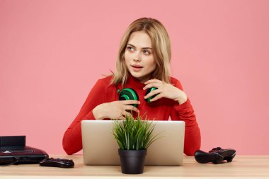 Kulaklık takan, dizüstü bilgisayarlı bir kadın masa başı eğlence sektöründe pembe arka planda oturuyor.