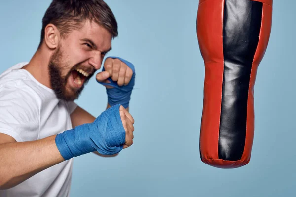 Un homme pratiquant un coup de poing sur un sac de boxe dans un t-shirt blanc sur un fond bleu — Photo