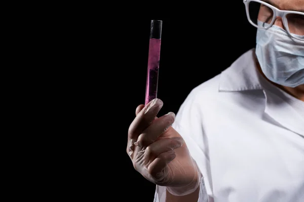 Laboratoriumassistent in een medisch masker en in een witte vacht met een vloeistof in een kolf en een chemische stof op een zwarte achtergrond — Stockfoto
