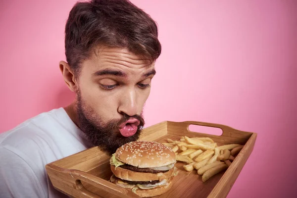 Man met een dienblad van voedsel in zijn hand hamburger frietjes en fast food calorie roze achtergrond portret close-up — Stockfoto