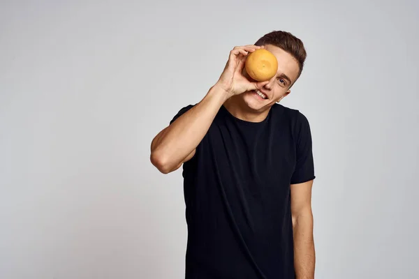 En kille med en orange i handen och i en svart T-shirt på en ljus bakgrund beskuren vy — Stockfoto