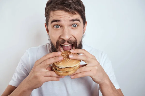 Egy férfi hamburgert eszik könnyű háttérrel fehér pólóban kivágott szemmel közelről éhség gyorsétterem — Stock Fotó