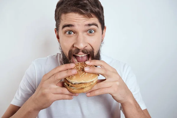 Seorang pria makan hamburger dengan latar belakang yang ringan dengan kaos putih dipotong memandang dari dekat kelaparan makanan cepat saji — Stok Foto