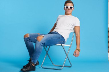 Kotlu, dizleri yırtık rahat bir adam mavi arka planda bir sandalyede oturuyor ve yüzünde uzun gözlüklerle oturuyor.