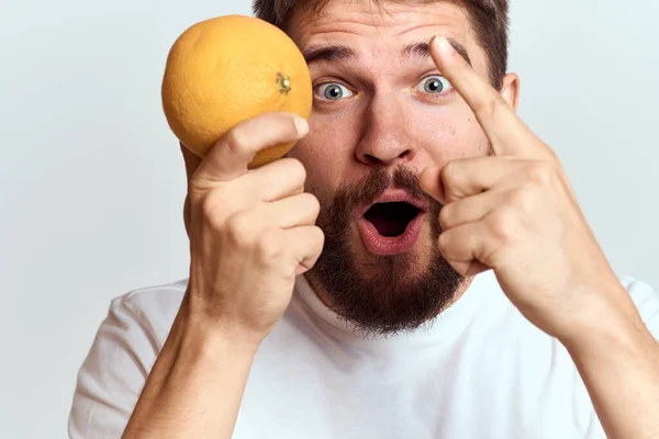 Un hombre con una naranja en una camiseta blanca sobre un fondo claro la dieta de una barba gruesa morena — Foto de Stock