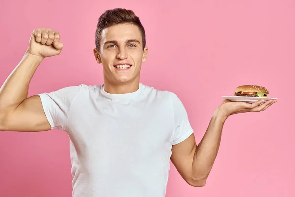 접시 위에 햄버거를 얹고 흰색 티셔츠 핑크 색 배경을 가진 감정적 인 남자 패스트푸드 칼로리의 전망이 흐트러지는 모습 — 스톡 사진