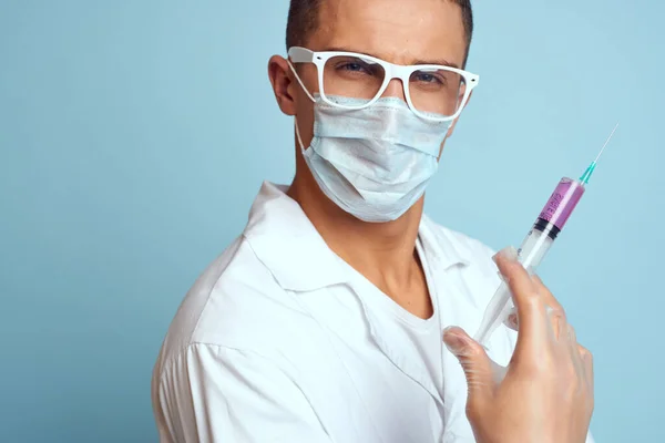 El médico sostiene una jeringa en la mano con un líquido rojo sobre un vestido médico de fondo azul y una máscara protectora. —  Fotos de Stock