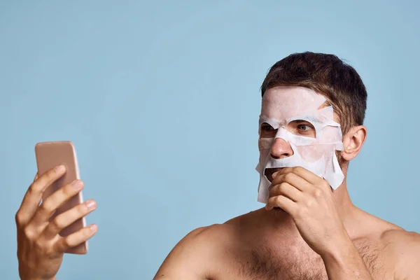 Um homem com uma máscara de limpeza em seu rosto examina-se em um espelho em um fundo azul — Fotografia de Stock