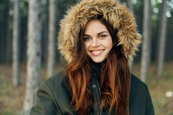 Mujer sonriente en una chaqueta con una capucha sobre un fondo de bosque —  Fotos de Stock