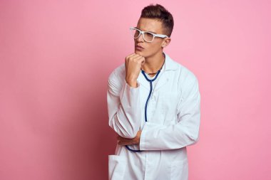 Pembe bir arka planda steteskop ve yüzünde gözlük olan tıbbi önlüklü bir erkek doktor.