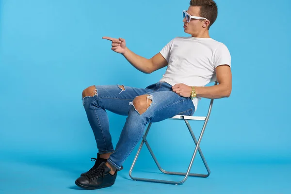 Ontspannen man in jeans met gescheurde knieën zit op een stoel binnen op een blauwe achtergrond met een full-length bril op zijn gezicht — Stockfoto