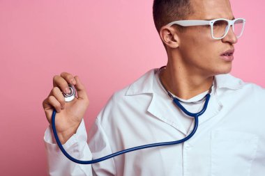 Pembe bir arka planda steteskop ve yüzünde gözlük olan tıbbi önlüklü bir erkek doktor.