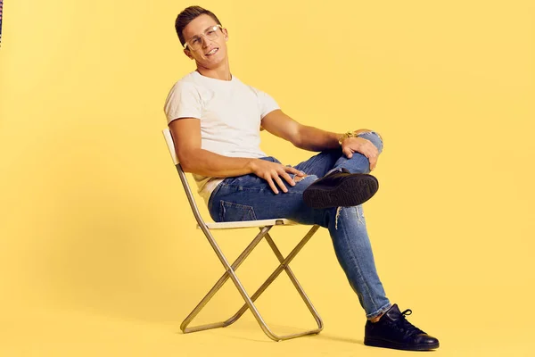 可爱的男人坐在椅子上，白色T恤衫牛仔裤，现代风格的黄色背景 — 图库照片